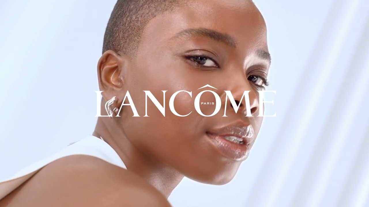 Modelka s krátkými vlasy se usmívá na kameru, logo Lancôme v popředí. Elegantní a moderní vzhled.